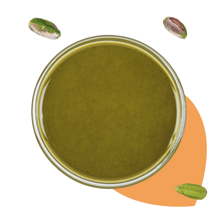 Foto vista dall'alto di un bicchiere di vetro pieno di pasta pura di pistacchio. Nella foto sono presenti anche dei pistacchi e un elemento astratto di colore arancione.